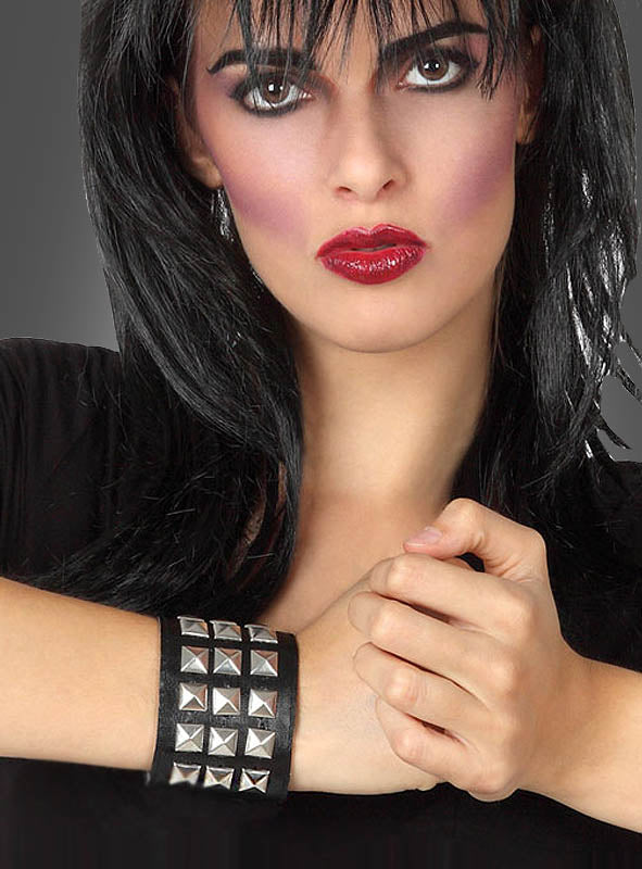 Eine Frau mit dunklem Haar und auffälligem Make-up trägt ein schwarzes Shirt und präsentiert das dreireihige Nietenarmband „Punk Rock“, während sie mit der anderen Hand ihr Handgelenk vor einem neutralen Hintergrund umklammert.