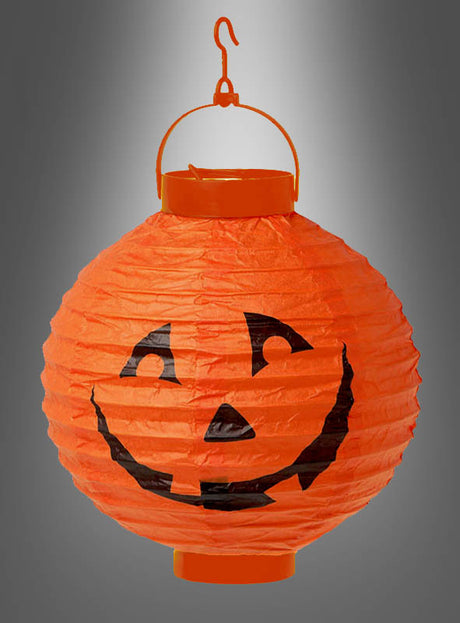 Kürbis-Lampion mit LED, 20 cm: Orangefarbener Papierlampion mit schwarzem Jack-o-Lantern-Gesicht, ideal als Halloween-Dekoration. Der Lampion hängt an einem Kunststoffhaken vor einem grauen Hintergrund mit Farbverlauf und sorgt dank integrierter LED für stimmungsvolles Licht.