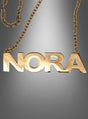 Deluxe-NORA-Kette für Pop-Duo: Eine goldene Metallkette mit einem Anhänger, auf dem „NORA“ in auffälligen Großbuchstaben zu sehen ist – die ideale Wahl für eine stilvolle 80er-Jahre-Mottoparty. Präsentiert wird sie vor einem eleganten grauen Farbverlauf.