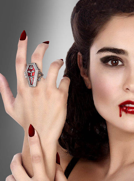Eine Frau mit Vampir-Make-up, rotem Lippenstift und Kunstblut trägt den Giftring in Sargform für Vampire mit einem Kreuz – ein perfekter Vampir-Schmuck und ein Gothic-Kostüm-Accessoire. Der Hintergrund verläuft von Grau nach Weiß.