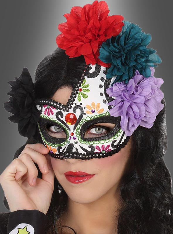 Eine Person, die die „Augenmaske Tag der Toten“ mit Blumenmustern und bunten Stoffblumen trägt und die Maske mit einer Hand vor einem grauen Hintergrund hält.
