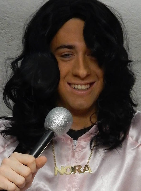 Eine Person mit langen dunklen Haaren, die eine „Deluxe NORA Kette für Pop Duo“ trägt und ein glitzerndes Mikrofon in der Hand hält, lächelt in die Kamera und ist bereit für eine 80er-Jahre-Mottoparty.