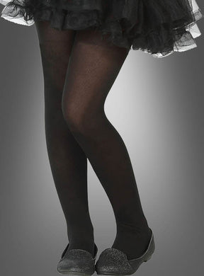 Eine Person trägt die Nylon-Strumpfhose „Kinder schwarz“, dazu einen schwarzen Rüschenrock und schwarze flache Schuhe, und steht mit überkreuzten Beinen vor einem grauen Hintergrund mit Farbverlauf.