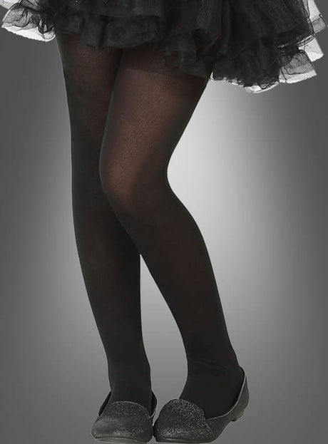Eine Person trägt die Nylon-Strumpfhose „Kinder schwarz“, dazu einen schwarzen Rüschenrock und schwarze flache Schuhe, und steht mit überkreuzten Beinen vor einem grauen Hintergrund mit Farbverlauf.