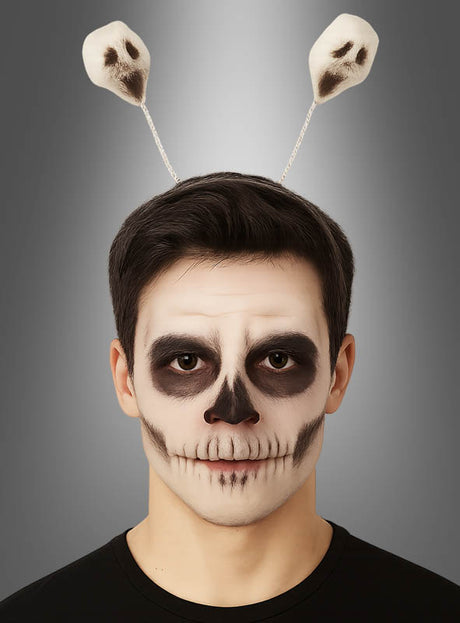 Eine Person mit einer Gesichtsbemalung, die einem Schädel ähnelt, und einem Halloween-Haarreif mit Geistern – einem Stirnband mit zwei auf Federn befestigten Geistern aus Schaumstoff – vor einem neutralen Hintergrund mit Farbverlauf.