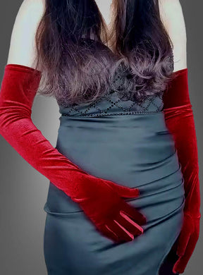 Eine Frau in einem schwarzen, ärmellosen Kleid mit Perlenverzierungen trägt die „Lange Handschuhe Samt rot“ als elegantes Accessoire. Sie steht vor einem grauen Hintergrund, wobei ihr Gesicht auf dem Bild nicht zu sehen ist.