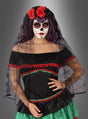 Vor einem grauen Hintergrund steht eine Person, die ein farbenfrohes „Dia de los Muertos“-Kostüm trägt, mit aufwendigem Zuckerschädel-Gesichts-Make-up, einem schwarzen Kleid mit roten und grünen Verzierungen, einem schwarzen Spitzenschleier und dem auffälligen „Señorita Blumenkranz mit Schleier“ als Kopfschmuck.
