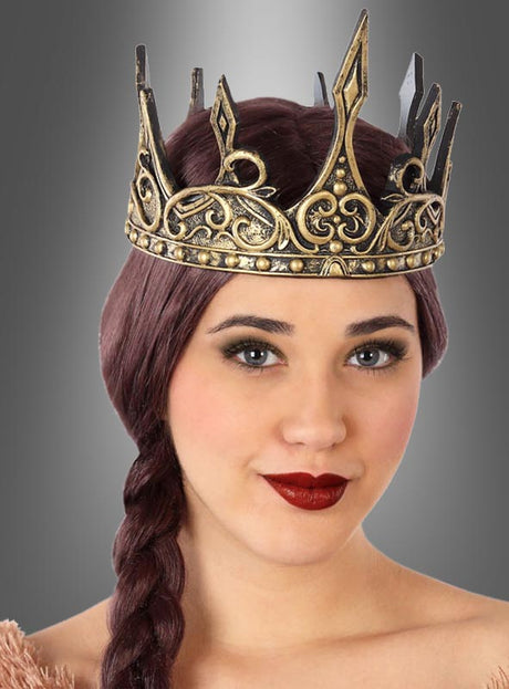 Frau mit langen, dunklen, geflochtenen Haaren, die eine verzierte Latexkrone „Königin Deluxe“ in Gold trägt, mit neutralem Gesichtsausdruck, vor einem grauen Hintergrund mit Farbverlauf.