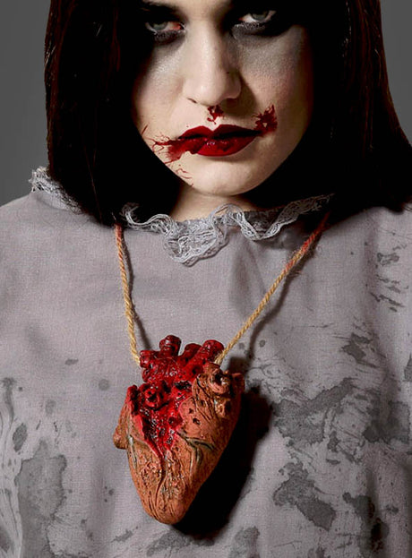 Eine Person mit auffälligem Make-up und blutroten Lippen trägt die Halloween-Halskette „Blutiges Herz Deko“ – eine Halskette mit einem realistisch gestalteten, blutigen Herz an einer Kordel – über einem grauen, fleckigen Shirt, was eine gewagte Wahl an Halloween-Schmuck darstellt.