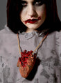 Eine Person mit auffälligem Make-up und blutroten Lippen trägt die Halloween-Halskette „Blutiges Herz Deko“ – eine Halskette mit einem realistisch gestalteten, blutigen Herz an einer Kordel – über einem grauen, fleckigen Shirt, was eine gewagte Wahl an Halloween-Schmuck darstellt.