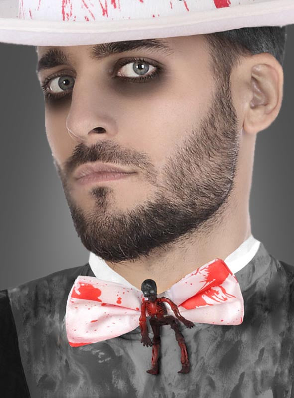 Ein Mann mit dunklem Augen-Make-up und Bart trägt einen weißen Hut, der mit der „Blutige Skelett Fliege“ als auffälliges Halloween-Accessoire verziert ist; an der Fliege ist eine kleine Zombie-Figur befestigt.