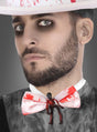 Ein Mann mit dunklem Augen-Make-up und Bart trägt einen weißen Hut, der mit der „Blutige Skelett Fliege“ als auffälliges Halloween-Accessoire verziert ist; an der Fliege ist eine kleine Zombie-Figur befestigt.