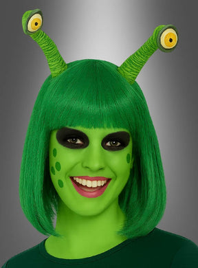 Eine Person mit grüner Haut, grüner Perücke und zwei „Alien-Augen“, die mit einem Haargummi am Kopf befestigt sind, trägt schwarzes Augen-Make-up und grüne Flecken auf den Wangen und lächelt in die Kamera. Dieser kreative Look wird durch dieses einzigartige Accessoire abgerundet, das für einen außerirdischen Look sorgt.