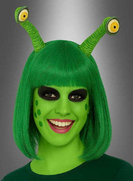 Eine Person mit grüner Haut, grüner Perücke und zwei „Alien-Augen“, die mit einem Haargummi am Kopf befestigt sind, trägt schwarzes Augen-Make-up und grüne Flecken auf den Wangen und lächelt in die Kamera. Dieser kreative Look wird durch dieses einzigartige Accessoire abgerundet, das für einen außerirdischen Look sorgt.