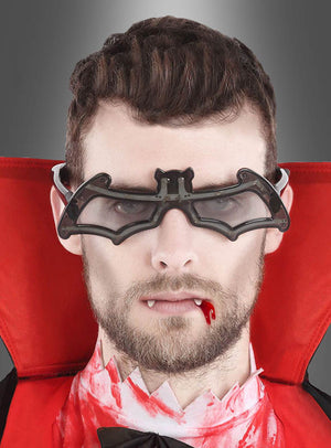 Ein als Vampir verkleideter Mann, der eine fledermausförmige Brille mit LED-Beleuchtung trägt, mit künstlichen Reißzähnen, Kunstblut am Mund und einem rot gefütterten Umhang, blickt mit ausdruckslosem Gesicht direkt in die Kamera.