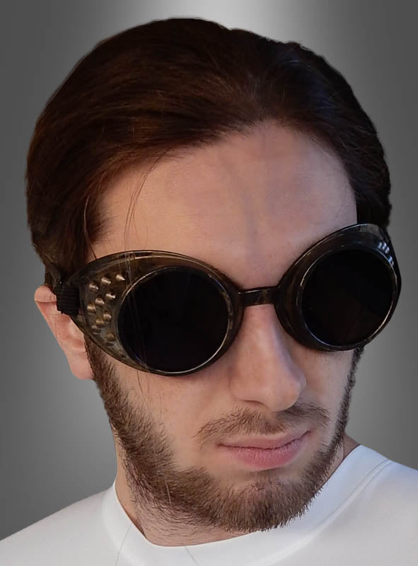 Ein Mann mit braunen Haaren und Bart trägt eine große, runde Steampunk-Brille und ein weißes Hemd vor einem grauen Hintergrund mit Farbverlauf.