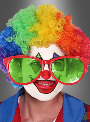 Eine Person mit einer Regenbogenperücke, einer roten Nase und einer übergroßen roten „Riesenbrille für Clowns“-Sonnenbrille, mit weißer Gesichtsbemalung und buntem Clown-Make-up, gekleidet in ein blau-rotes Kostüm, lächelt in die Kamera – bereit, auf jeder Schlagerparty für gute Laune zu sorgen.