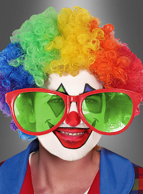 Eine Person mit einer Regenbogenperücke, einer roten Nase und einer übergroßen roten „Riesenbrille für Clowns“-Sonnenbrille, mit weißer Gesichtsbemalung und buntem Clown-Make-up, gekleidet in ein blau-rotes Kostüm, lächelt in die Kamera – bereit, auf jeder Schlagerparty für gute Laune zu sorgen.
