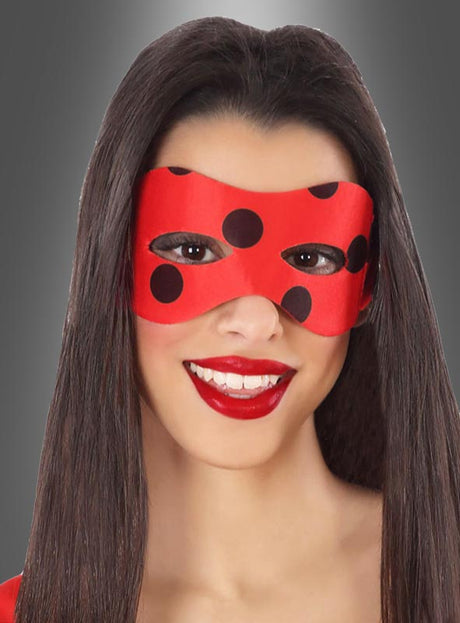 Eine Frau mit langen, glatten Haaren lächelt vor einem grauen Hintergrund mit Farbverlauf in die Kamera. Sie trägt die „Marienkäfer-Augenmaske“ – eine leuchtend rote Maske mit schwarzen Punkten – sowie roten Lippenstift. Diese einzigartige „Marienkäfer-Augenmaske“ verleiht ihrem Look eine verspielte Note.