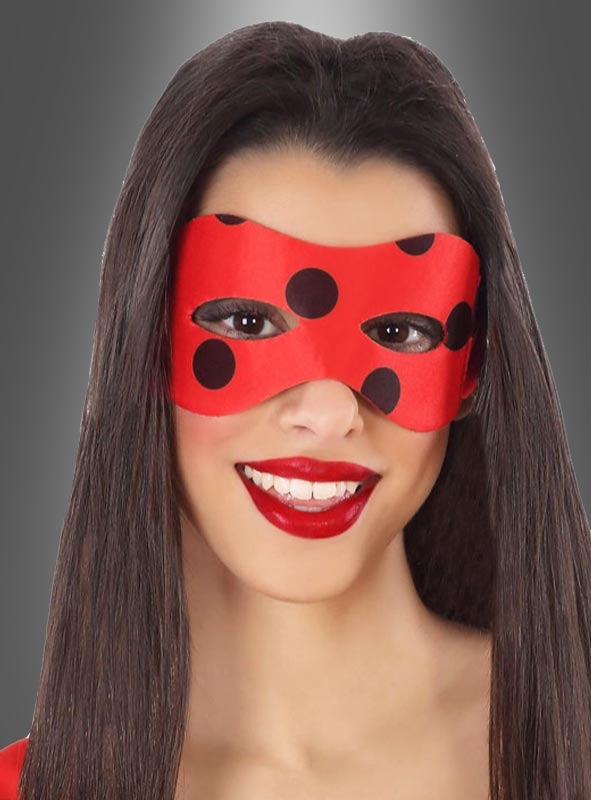 Eine Frau mit langen, glatten Haaren lächelt vor einem grauen Hintergrund mit Farbverlauf in die Kamera. Sie trägt die „Marienkäfer-Augenmaske“ – eine leuchtend rote Maske mit schwarzen Punkten – sowie roten Lippenstift. Diese einzigartige „Marienkäfer-Augenmaske“ verleiht ihrem Look eine verspielte Note.