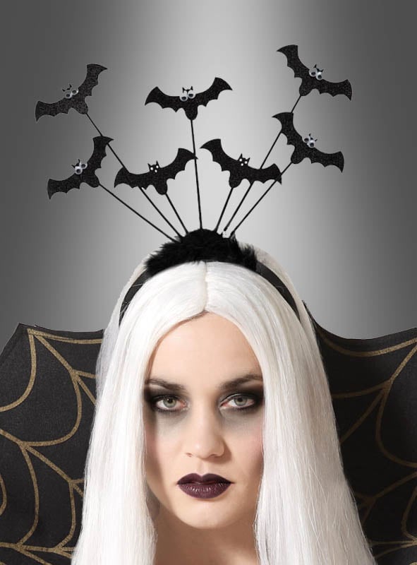 Eine Frau mit langen weißen Haaren trägt die „Fledermäuse auf Haarreif mit Wackelaugen“, ein verspieltes Halloween-Accessoire mit schwarzen Fledermäusen mit Wackelaugen an Drähten. Sie rundet ihr elegantes Vampir-Kostüm mit dramatischem Augen-Make-up, schwarzem Lippenstift und einem gemusterten schwarzen Umhang ab. Die Szene spielt sich vor einem grauen Hintergrund mit Farbverlauf ab.