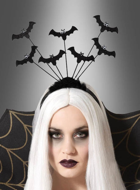 Eine Frau mit langen weißen Haaren trägt die „Fledermäuse auf Haarreif mit Wackelaugen“, ein verspieltes Halloween-Accessoire mit schwarzen Fledermäusen mit Wackelaugen an Drähten. Sie rundet ihr elegantes Vampir-Kostüm mit dramatischem Augen-Make-up, schwarzem Lippenstift und einem gemusterten schwarzen Umhang ab. Die Szene spielt sich vor einem grauen Hintergrund mit Farbverlauf ab.