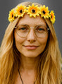 Eine Person mit langen blonden Haaren trägt eine runde Brille und einen gelben Sonnenblumen-Haarkranz für Hippies, was an einen klassischen Damenanzug erinnert, und blickt vor einem schlichten Hintergrund in die Kamera.