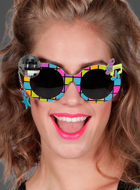 Eine Frau mit lockigem Haar trägt die farbenfrohe 70er-Jahre-Disco-Brille mit einem verspielten karierten Rahmen, der mit einer Discokugel, blauen Sternen und einer Musiknote verziert ist – das perfekte Accessoire, um deinen nächsten Disco-Kostüm-Look abzurunden. Sie lächelt strahlend und trägt leuchtend pinken Lippenstift.