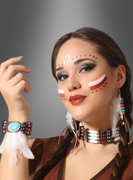 Eine Frau mit geflochtenen Haaren trägt das „Indianer Federschmuck“-Set (4-teilig), bestehend aus Federohrringen, Perlen-Schmuck und Gesichtsbemalung mit weißen und roten Linien und Punkten, während sie vor einem grauen Hintergrund posiert.