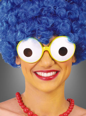 Eine Person mit einer großen blauen Lockenperücke, einer riesigen Comic-Brille mit lustigem Glotzaugen-Effekt, einer roten Perlenkette und rotem Lippenstift, die in die Kamera lächelt.