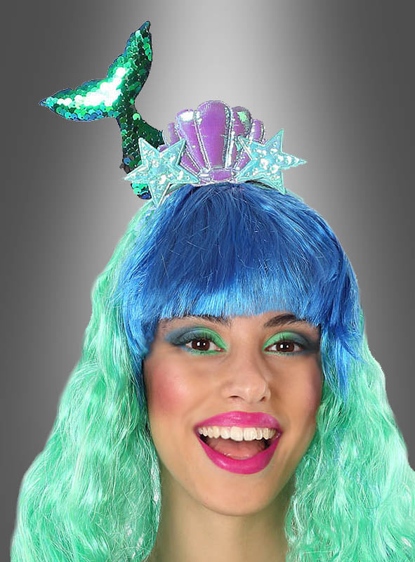 Eine Person mit einer blau-grünen Perücke lächelt vor einem grauen Hintergrund mit Farbverlauf und trägt den Haarschmuck „Meerjungfrau“ – verziert mit einer violetten Muschel, zwei Sternen und einer grünen, glitzernden Meerjungfrauenflosse – das perfekte Accessoire, um jedes Meerjungfrauenkostüm zu vervollständigen.