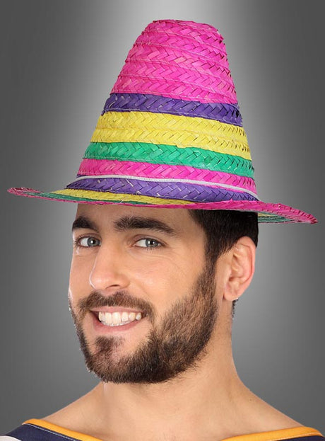 Ein bärtiger Mann lächelt, während er den „Kleiner Mexikaner Hut“-Hut trägt, einen hohen, farbenfrohen, geflochtenen Hut mit Streifen in Rosa, Gelb, Grün und Lila.