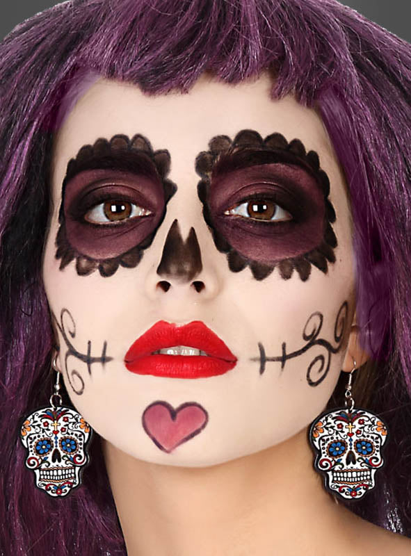 Eine Person mit lila Haaren und „Tag der Toten“-Gesichtsbemalung, bestehend aus schwarzen Augenringen, einem Herz am Kinn und auffälligen „Tag der Toten“-Totenkopf-Ohrringen – das perfekte Halloween-Kostüm-Accessoire, um den Look zu vervollständigen.