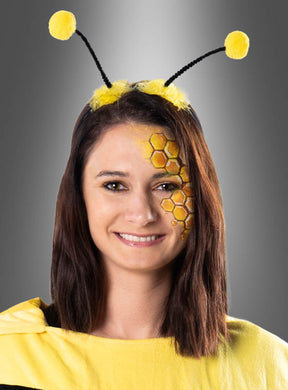 Eine Frau in einem Bienenkostüm mit einem Haarreif in Form von Bienenantennen mit gelben Pompons und einer Waben-Gesichtsbemalung auf einer Wange lächelt vor einem grauen Hintergrund in die Kamera.