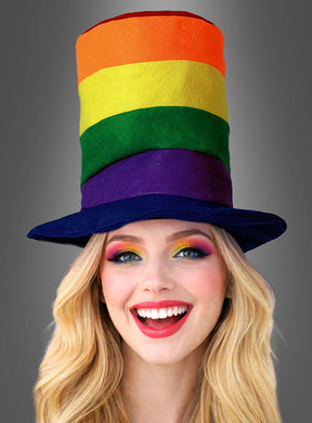 Eine lächelnde Person mit langen blonden Haaren trägt den „Regenbogen-Stoffzylinder“, einen hohen Stoffhut mit Regenbogenstreifen, der vor einem einfarbigen grauen Hintergrund abgebildet ist.