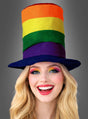 Eine lächelnde Person mit langen blonden Haaren trägt den „Regenbogen-Stoffzylinder“, einen hohen Stoffhut mit Regenbogenstreifen, der vor einem einfarbigen grauen Hintergrund abgebildet ist.