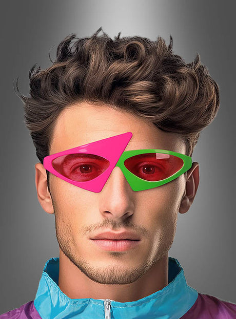 Ein Mann mit kurzen braunen Haaren trägt eine asymmetrische 80er-Jahre-Brille – zwei ungleiche geometrische Sonnenbrillengläser, eines in Rosa und eines in Grün – sowie eine lila-blaue Jacke vor einem grauen Hintergrund mit Farbverlauf.