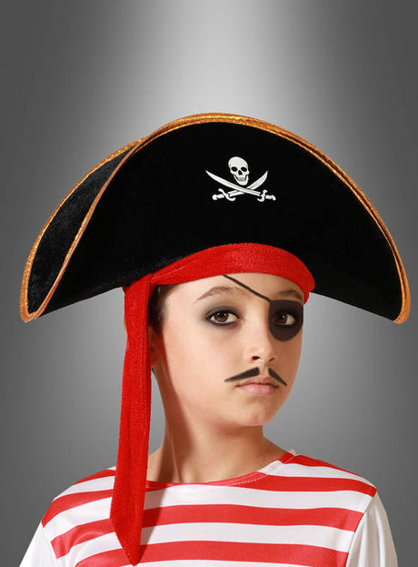 Ein Kind in einem rot-weiß gestreiften Hemd trägt den „Piratenhut für Kinder“ – einen schwarzen Hut mit Totenkopf, ergänzt durch ein rotes Bandana, schwarzes Augen-Make-up und einen aufgemalten Schnurrbart –, vor einem grauen Hintergrund mit Farbverlauf – das perfekte Piratenkostüm für jedes fantasievolle Abenteuer.