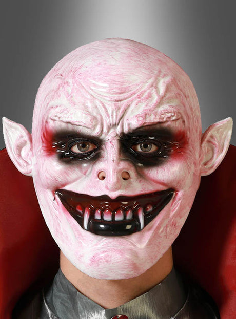 Eine Person, die die „Vampir-Maske für Erwachsene“ trägt – mit blassrosa Haut, spitzen Ohren, übertrieben großen Zähnen und auffälligem schwarz-rotem Make-up um die Augen –, steht vor einem grauen Hintergrund mit Farbverlauf.
