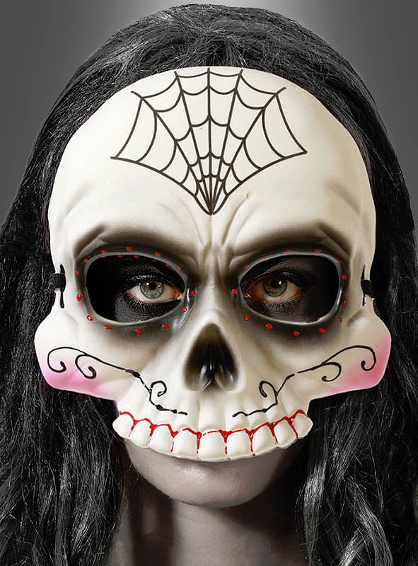 Eine Person, die die „Tag der Toten“-Halbmaske „Totenkopf“ trägt – eine weiße Maske mit schwarzen Augenlöchern, roten Akzenten, einem Spinnennetzmuster auf der Stirn und rosa Wangen. Dunkles Haar umrahmt die Maske.