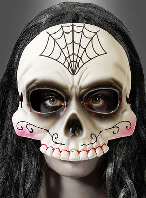 Eine Person, die die „Tag der Toten“-Halbmaske „Totenkopf“ trägt – eine weiße Maske mit schwarzen Augenlöchern, roten Akzenten, einem Spinnennetzmuster auf der Stirn und rosa Wangen. Dunkles Haar umrahmt die Maske.