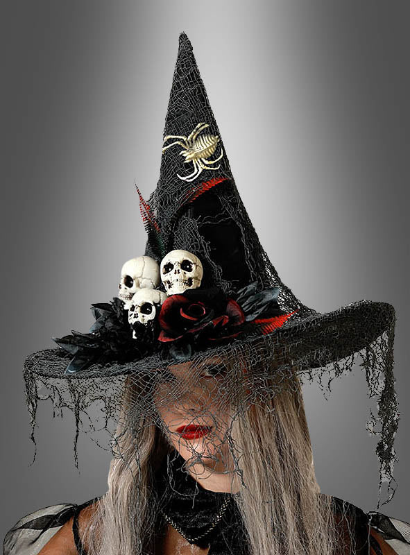 Eine Person, die den „Hexenhut mit Totenköpfen und Spinnen“ trägt – einen schwarzen Hexenhut, verziert mit Totenkopfmotiven, einer Spinne, einem roten Band, schwarzer Spitze und Kunstrosen – vor einem grauen Hintergrund mit Farbverlauf.