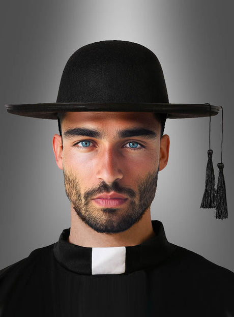Ein Mann mit blauen Augen und Bart trägt den schwarzen, breitkrempigen „Don Camillo“-Hut für Priester, der mit Quasten und einem Priesterkragen verziert ist, und erinnert vor einem grauen Hintergrund mit Farbverlauf an einen italienischen Pfarrer.