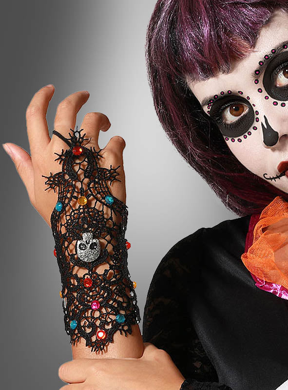 Eine Person mit lila Haaren und „Day of the Dead“-Make-up präsentiert die „Tag der Toten“-Armstulpe aus Spitze – einen schwarzen Spitzenhandschuh, verziert mit bunten Perlen und einem Totenkopf-Anhänger – und streckt ihre Hand vor einem grauen Hintergrund in die Höhe; dieses elegante Accessoire mit aufwendigen Spitzendetails eignet sich perfekt für jedes „Día de los Muertos“-Kostüm.