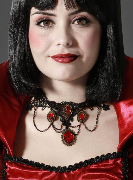 Eine Frau in einem auffälligen rot-schwarzen Kostüm mit dramatischem Kragen trägt die „Vampir Collier Kette“, eine faszinierende Halskette mit roten Edelsteinen. Ihr glatter schwarzer Bob und der kräftige rote Lippenstift unterstreichen den Look, während sie vor einem schlichten Hintergrund in die Kamera blickt – eine ideale Kulisse für Gothic-Schmuck oder als bezaubernde Halloween-Halskette.