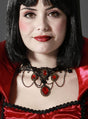 Eine Frau in einem auffälligen rot-schwarzen Kostüm mit dramatischem Kragen trägt die „Vampir Collier Kette“, eine faszinierende Halskette mit roten Edelsteinen. Ihr glatter schwarzer Bob und der kräftige rote Lippenstift unterstreichen den Look, während sie vor einem schlichten Hintergrund in die Kamera blickt – eine ideale Kulisse für Gothic-Schmuck oder als bezaubernde Halloween-Halskette.