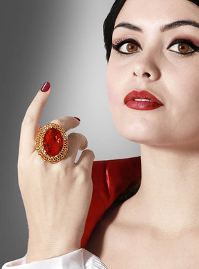 Eine Frau mit kurzen dunklen Haaren und rotem Lippenstift trägt den auffälligen Ring „Roter Rubin Ring Vampirin“ mit einem markanten roten Edelstein, während sie ihre gepflegte Hand mit den rot lackierten Nägeln an ihr Gesicht hält. Sie ist in ein auffälliges rot-weißes Outfit gekleidet.