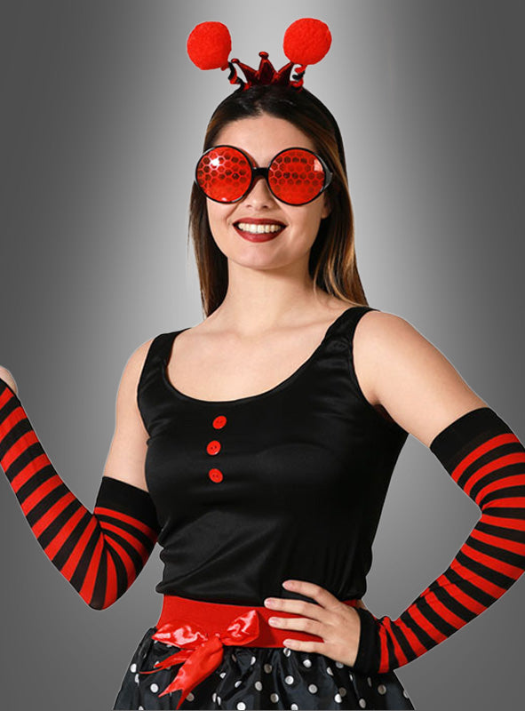 Eine Frau im Marienkäfer-Kostüm-Set, bestehend aus rot-schwarz gestreiften Handschuhen, roten Knöpfen, einem gepunkteten Rock, einer großen roten Brille und einem Stirnband mit Fühlern – eine auffällige Karnevalsverkleidung –, hebt sich vor einem grauen Hintergrund deutlich ab.