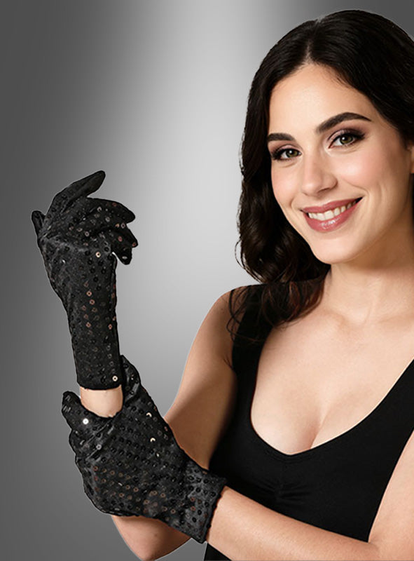 Eine Frau mit dunklem Haar lächelt, während sie „Kurze Pailletten-Handschuhe schwarz – schwarze Damenhandschuhe“ anzieht; kombiniert mit ihrem schwarzen, ärmellosen Oberteil und vor einem grauen Hintergrund mit Farbverlauf posierend, kreiert sie einen schicken Look, der an ein Kostüm-Accessoire aus den 20er Jahren erinnert.