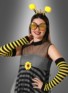 Eine Frau in einem Bienenkostüm – bestehend aus dem 4-teiligen Bienenkostüm-Set mit gestreiften Armstulpen, einer runden Sonnenbrille im Wabenmuster, einem Blumenakzent auf ihrem Kleid und einem Stirnband mit Fühlern – posiert vor einem grauen Hintergrund, um dieses auffällige Bienenkostüm-Zubehör für Damen zu präsentieren.